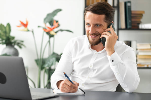 istockphoto-1320889536-612x612 Successful caucasian businessman talking by smartphone with somebody while working laptop in the office. Executive young businessman talking on mobile phone while sitting at work desk